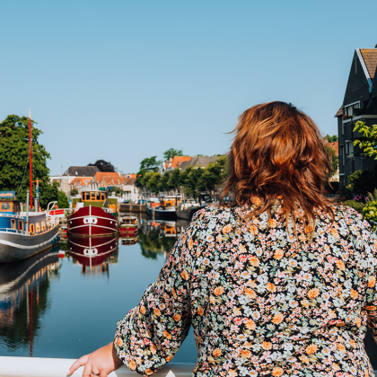 Vrouw leunt op brugreling en kijkt uit over water waarop boten aangemeerd liggen.